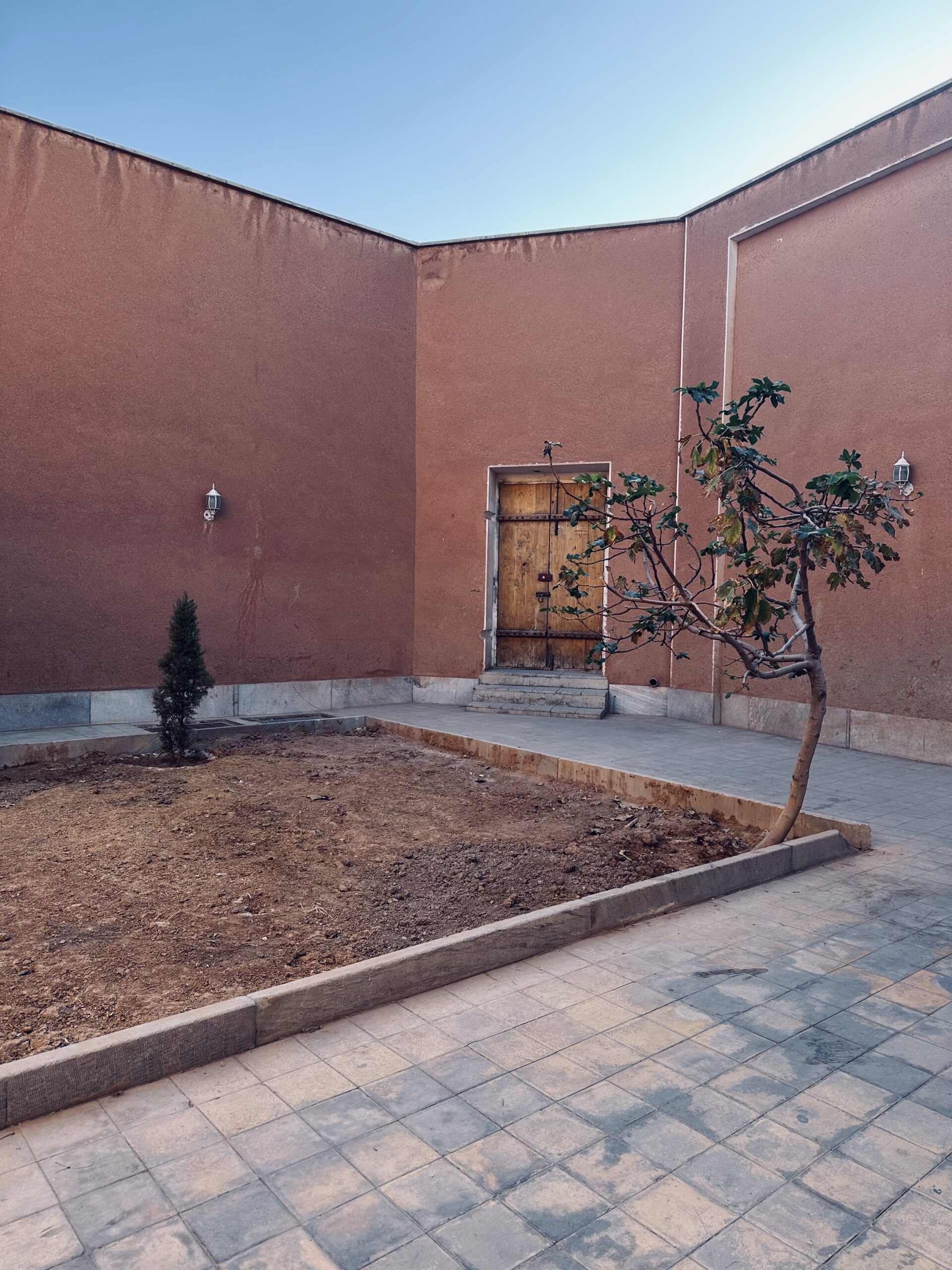 Quiet courtyard of Vank Cathedral in Isfahan with adobe-colored walls, a wooden door, a small tree, and clear blue sky overhead.