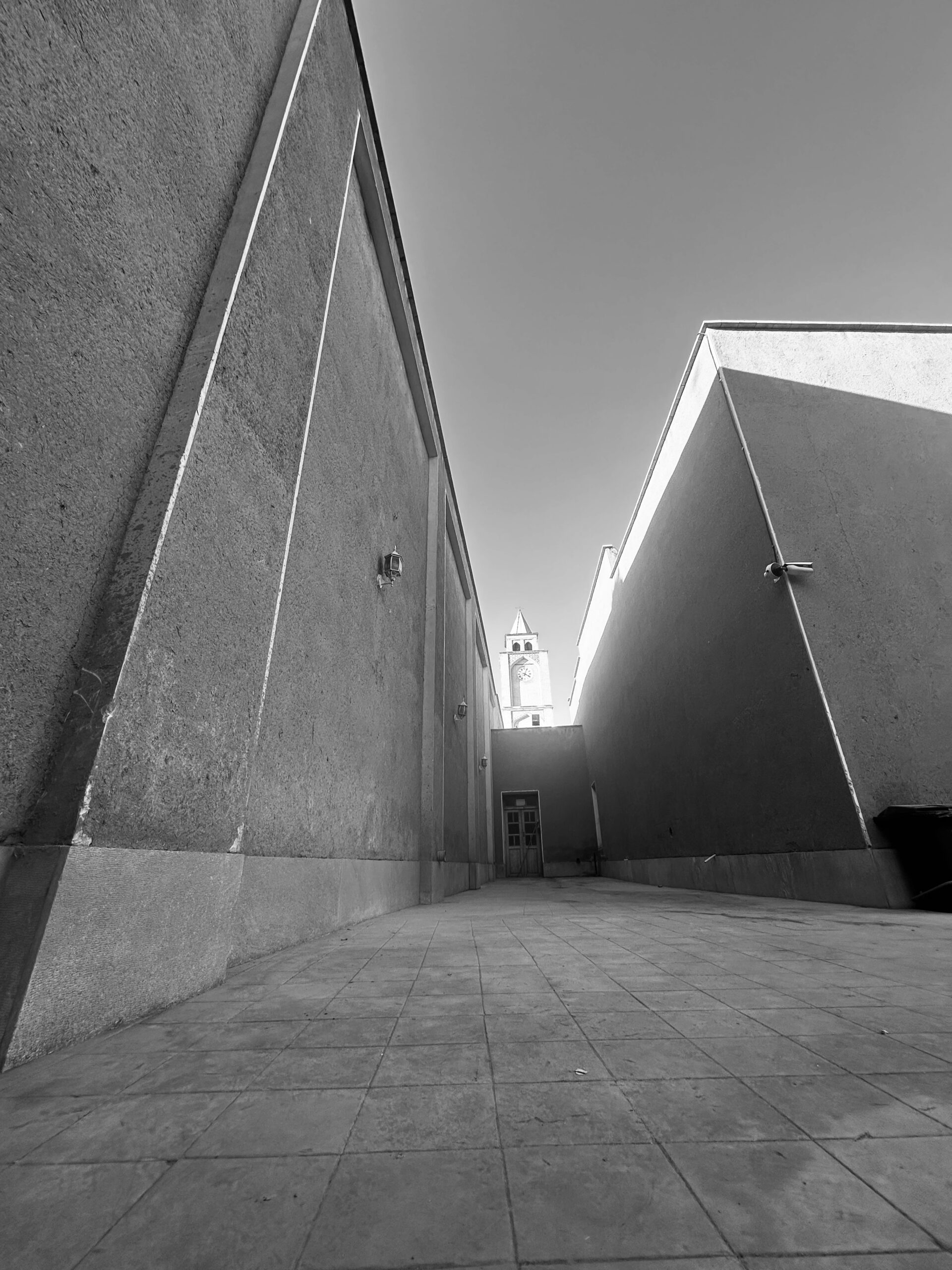 Black and white, low-angle shot of a narrow alleyway or courtyard leading toward the base of a clock tower, likely Vank Cathedral's bell tower, framed by two tall, plain, untextured walls on the left and right.